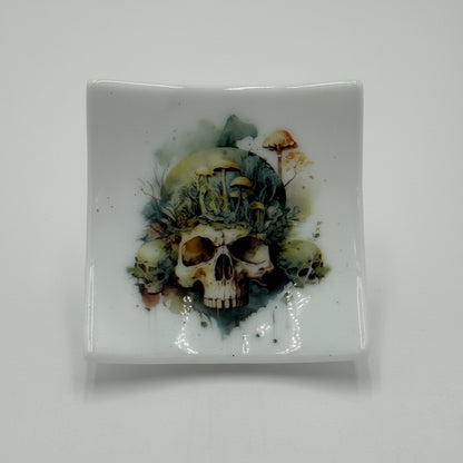 A square white ceramic dish featuring a detailed graphic of skulls and foliage in earthy green, brown, and black tones, displayed on a small clear stand against a plain white background.