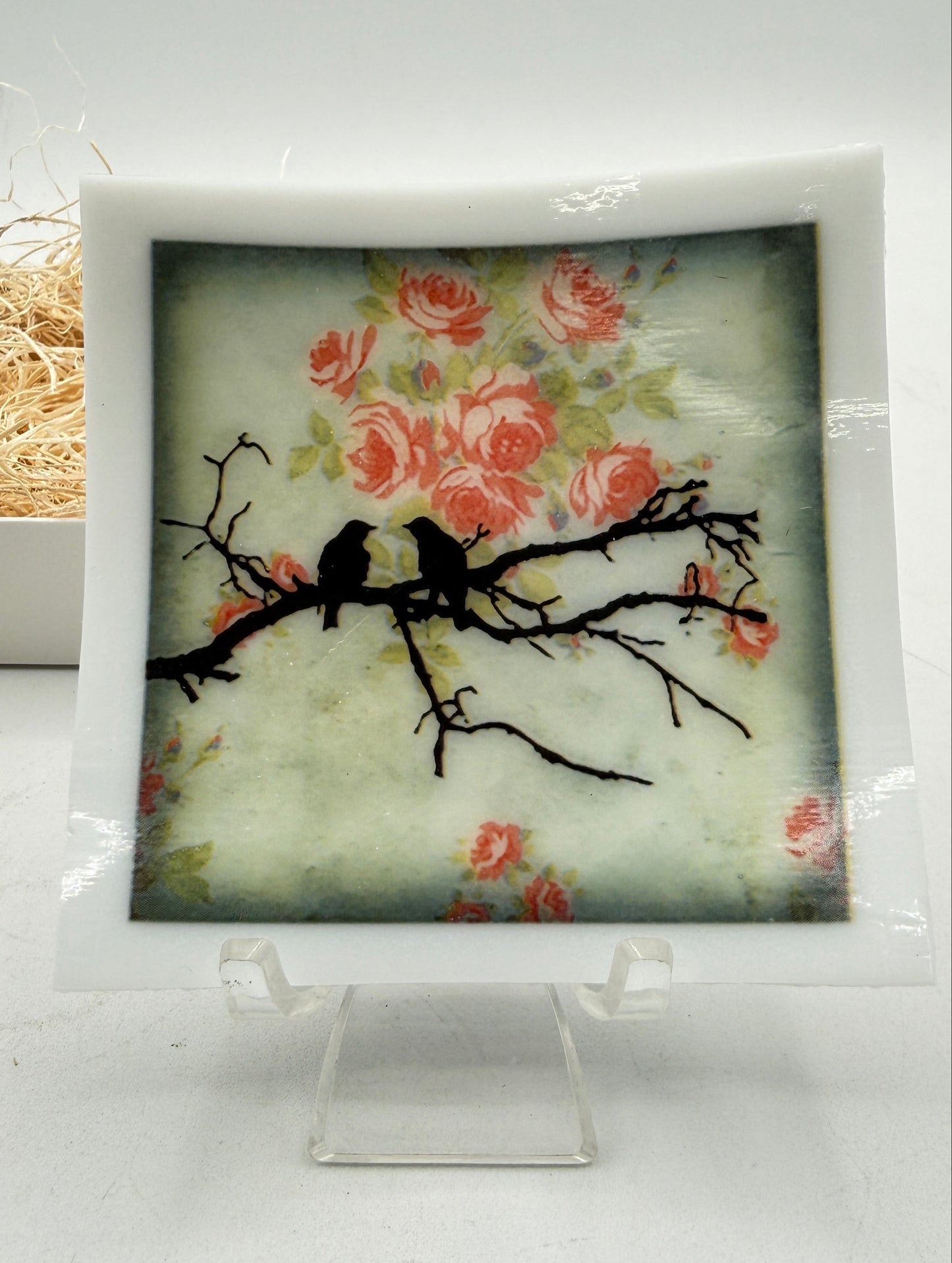 A square decorative plaque or tile with a white border, featuring a painted floral scene with pink roses, green leaves, two black bird silhouettes on a branch, and a soft blue-green background. The plaque is displayed on a clear acrylic stand against a white backdrop.