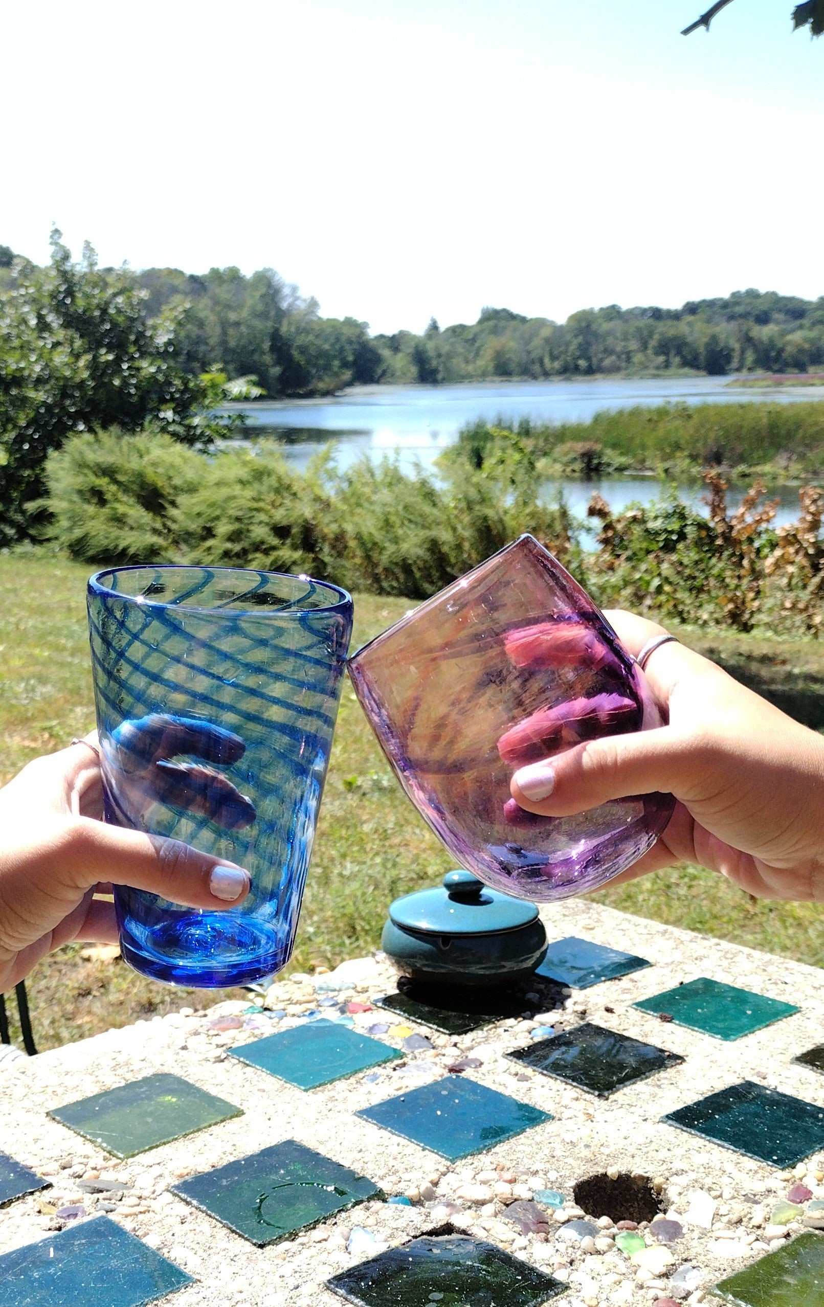 Couple clinking colorful hand-blown glasses outdoors by a lake for Date Night Glassblowing class