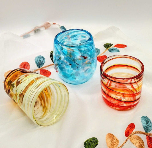 Three colorful handmade drinking glasses in yellow, blue, and red with artistic swirl patterns on a white fabric with multicolored leaf designs.