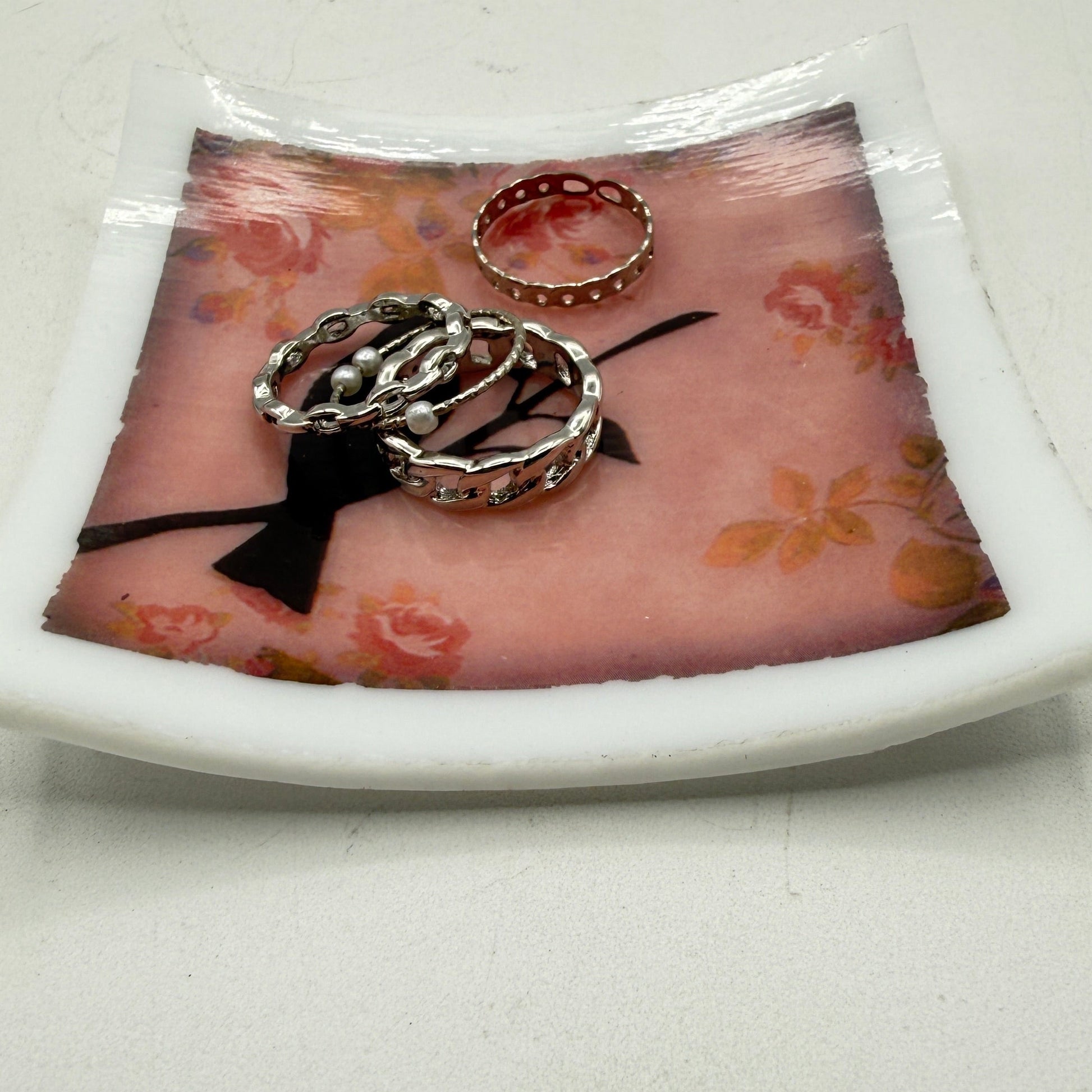 A white ceramic ring tray with a pink floral print and a black ribbon bow, holding three rings on a light gray surface.