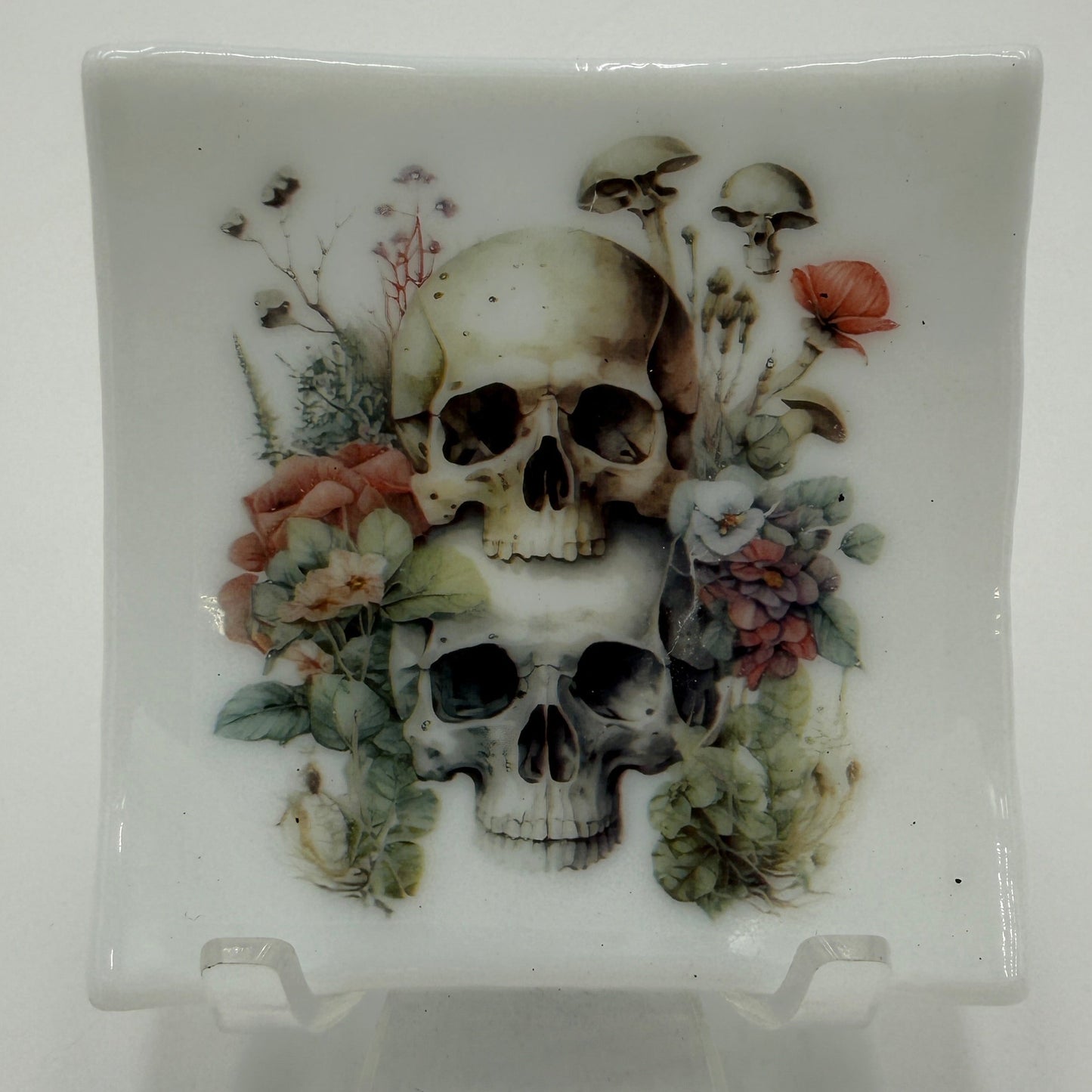 A square white ceramic dish with a glossy finish, featuring a detailed illustration of two human skulls surrounded by dried flowers, leaves, and mushrooms. The dish is displayed on a clear acrylic stand against a plain white background.