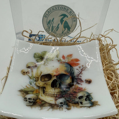 A square white ceramic plate with rounded corners, featuring a detailed skull illustration decorated with autumn leaves and small skulls. The plate is displayed inside a white gift box filled with brown packing paper, with a circular 'Blackstone River' logo card visible behind it.