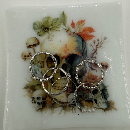 Three silver rings with clear crystal-like accents are displayed on a square white ceramic plate featuring a gothic illustration of skulls, flowers, and mushrooms.