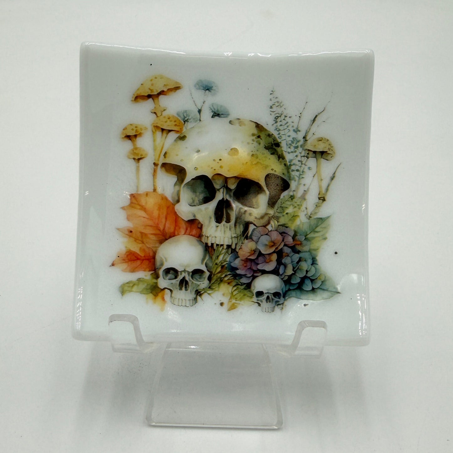 A square white ceramic plate with a hand-painted illustration of skulls, mushrooms, and autumn leaves, displayed on a clear plastic stand against a white background.