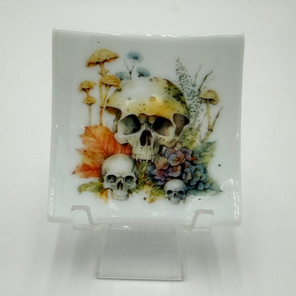 A square white ceramic plate with a hand-painted illustration of skulls, mushrooms, and autumn leaves, displayed on a clear plastic stand against a white background.