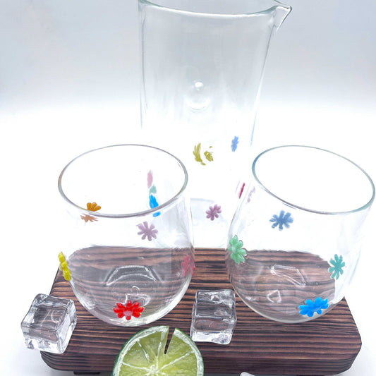 Hand blown stemless wine glasses with colorful retro flower designs and matching clear pitcher on wooden tray with ice cubes and lime wedge