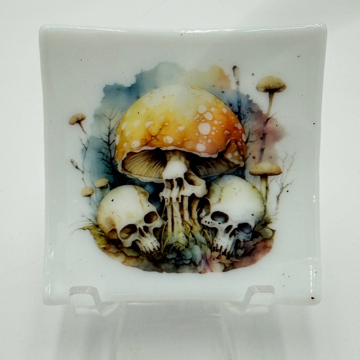 A square white ceramic plate with a detailed illustration of skulls and a large orange-brown mushroom on the front, displayed on a clear acrylic stand against a white background.