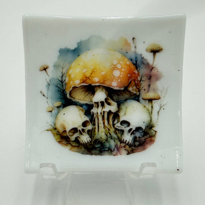 A square white ceramic plate with a detailed illustration of skulls and a large orange-brown mushroom on the front, displayed on a clear acrylic stand against a white background.