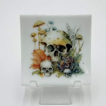 A square white ceramic plate with a hand-painted illustration of skulls, mushrooms, and autumn leaves, displayed on a clear acrylic stand against a white background.