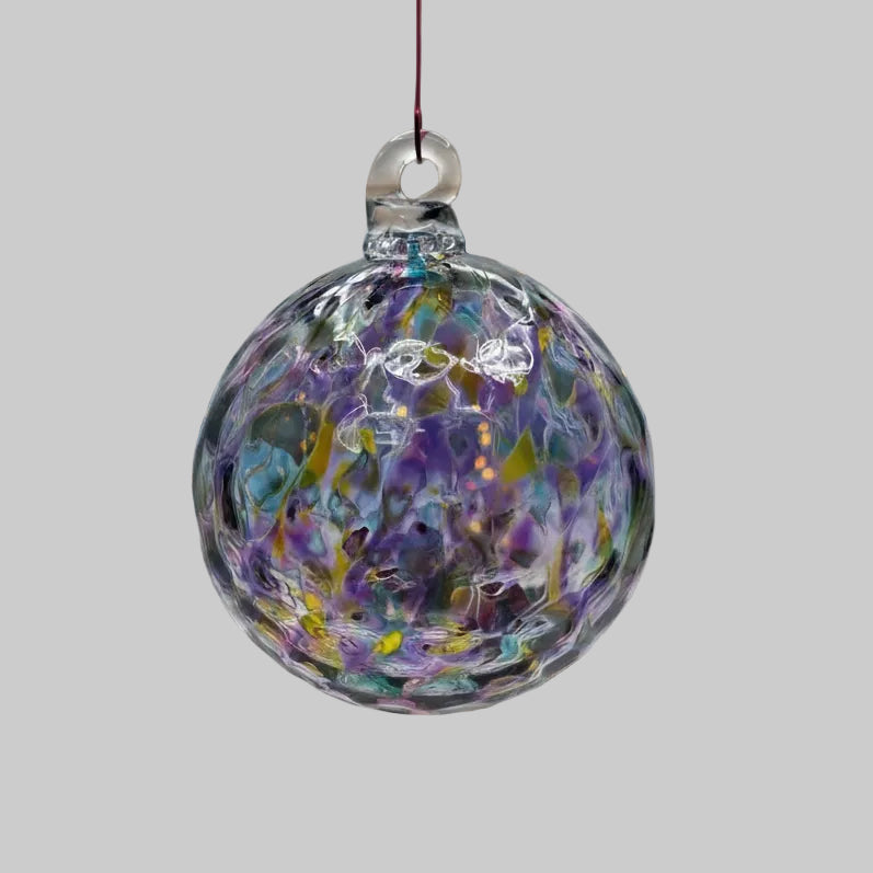 A hand-blown glass Christmas ornament with a clear glass body containing colorful swirls of purple, blue, yellow, and pink, suspended by a thin red string against a neutral gray background.