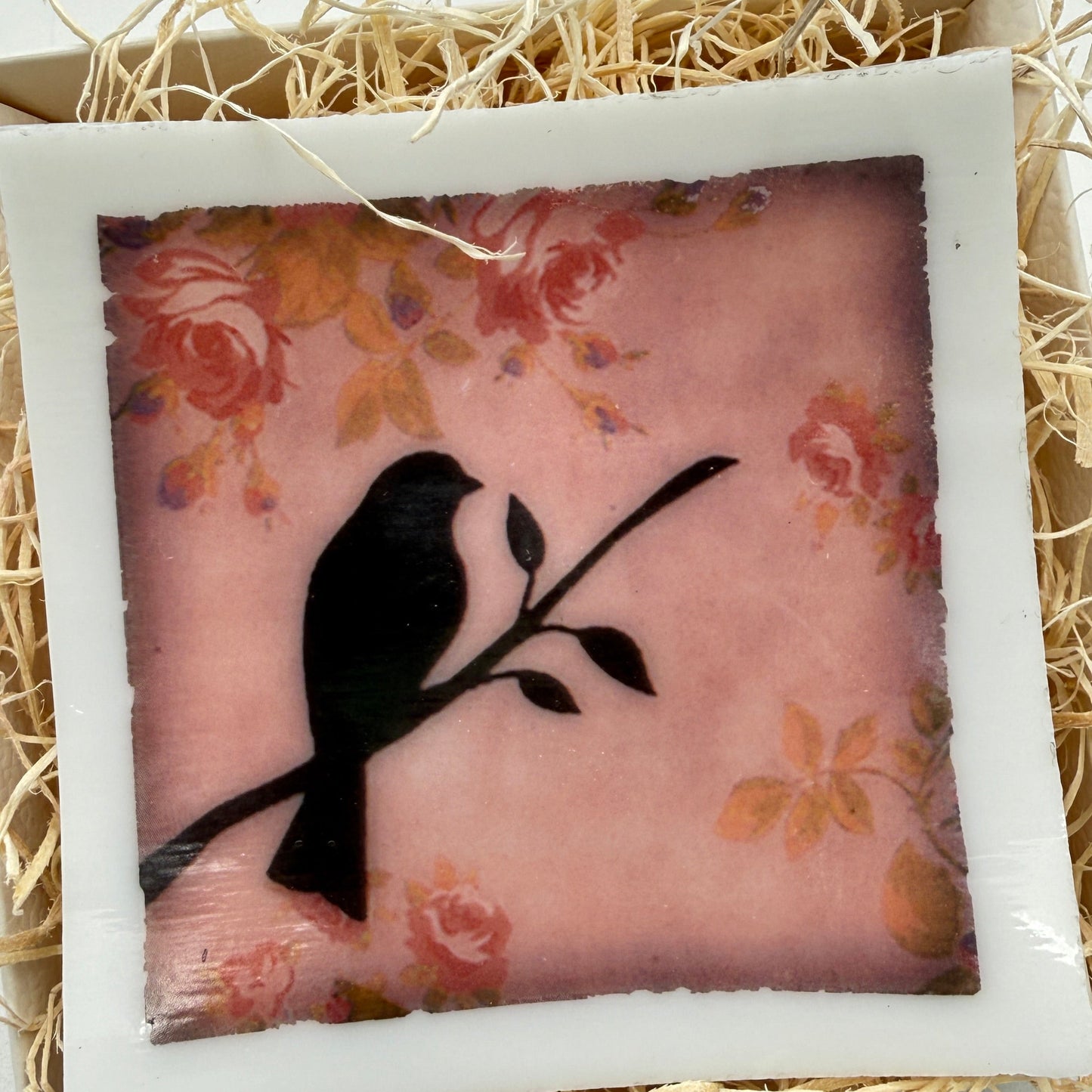 A square decorative plaque or tile with a white border, featuring a black bird silhouette perched on a branch over a pink floral background, displayed in a box filled with shredded straw.