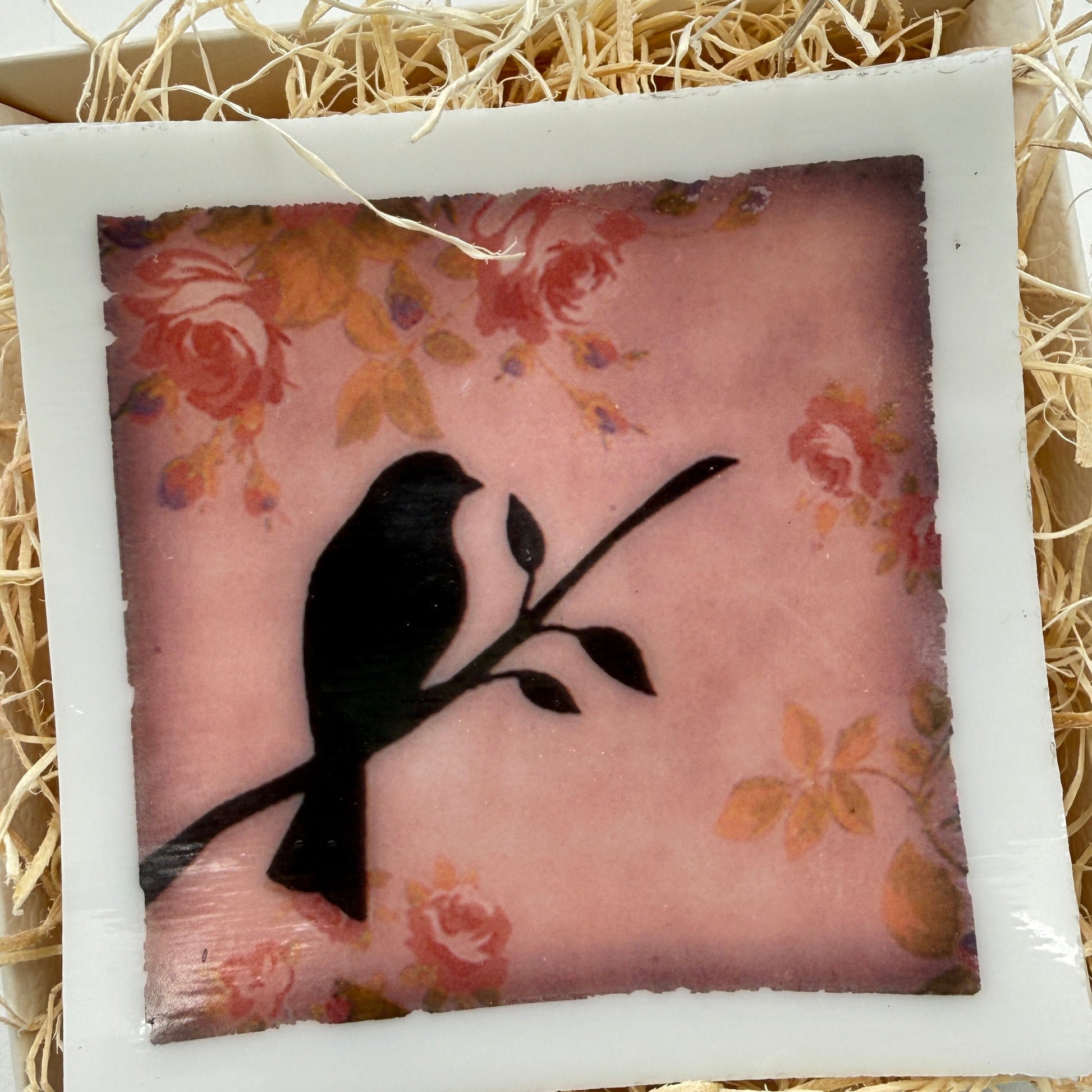 A square decorative plaque or tile with a white border, featuring a black bird silhouette perched on a branch over a pink floral background, displayed in a box filled with shredded straw.