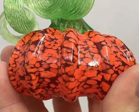 Handcrafted glass mini pumpkin with orange and black speckled design and green curly stem held in hands
