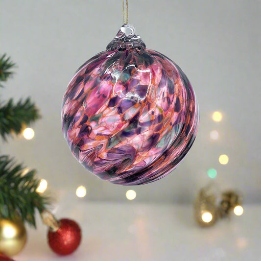 Colorful blown glass ornament with swirling pink, purple, and orange hues, displayed on a clear stand with festive silver and red garland nearby
