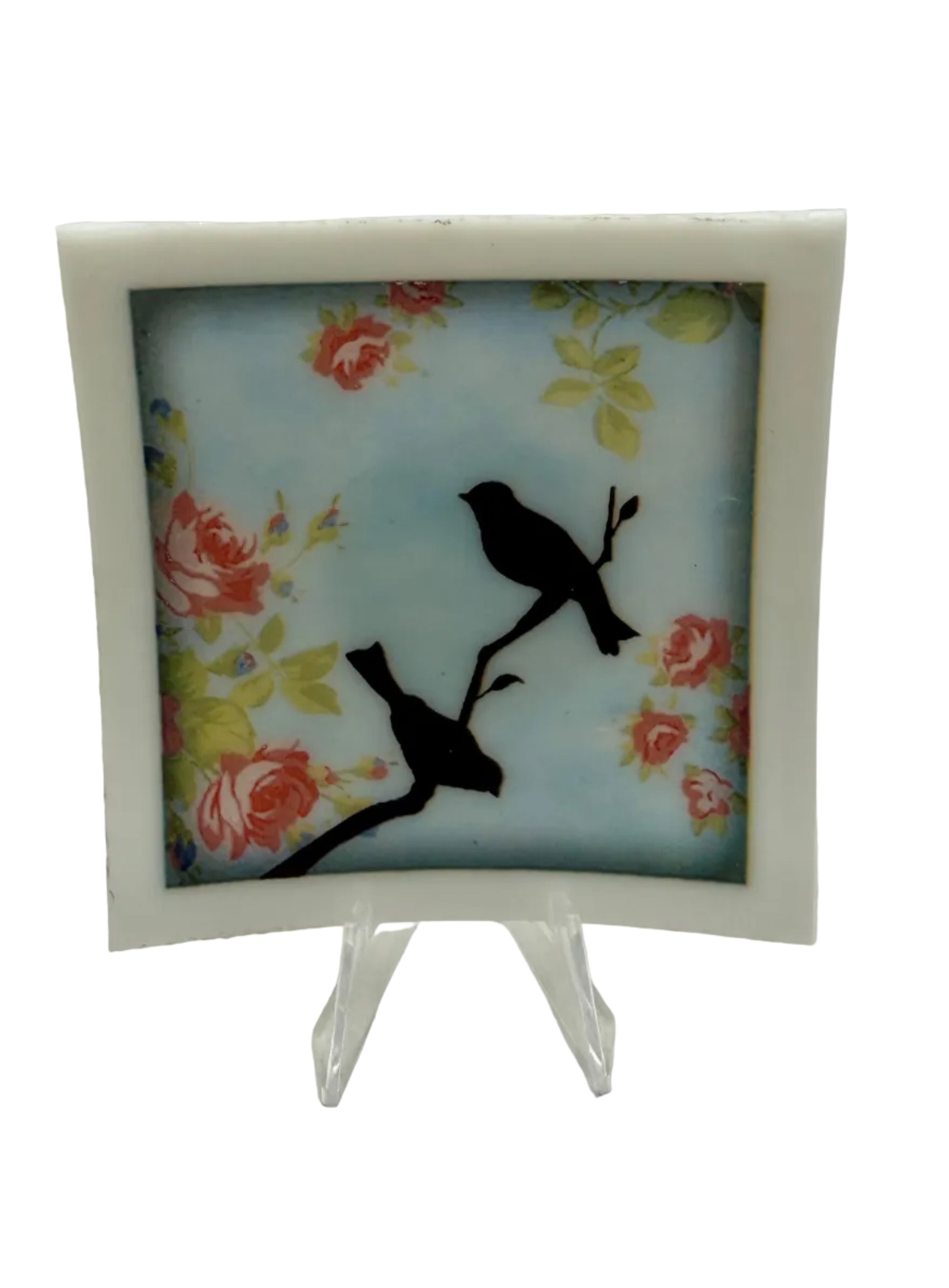 A square decorative art tile with a white border, featuring two black bird silhouettes perched on branches against a light blue background with red and yellow floral motifs, displayed on a clear stand.