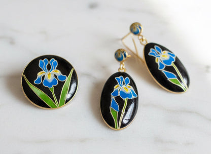 A close-up of a hand holding a small oval black stone or cabochon decorated with a blue and yellow flower and green leaves, with a gold band ring on the finger.