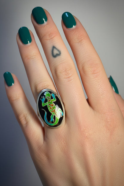 A close-up of a hand wearing a large silver ring with a black oval face featuring a colorful green and blue lizard design.