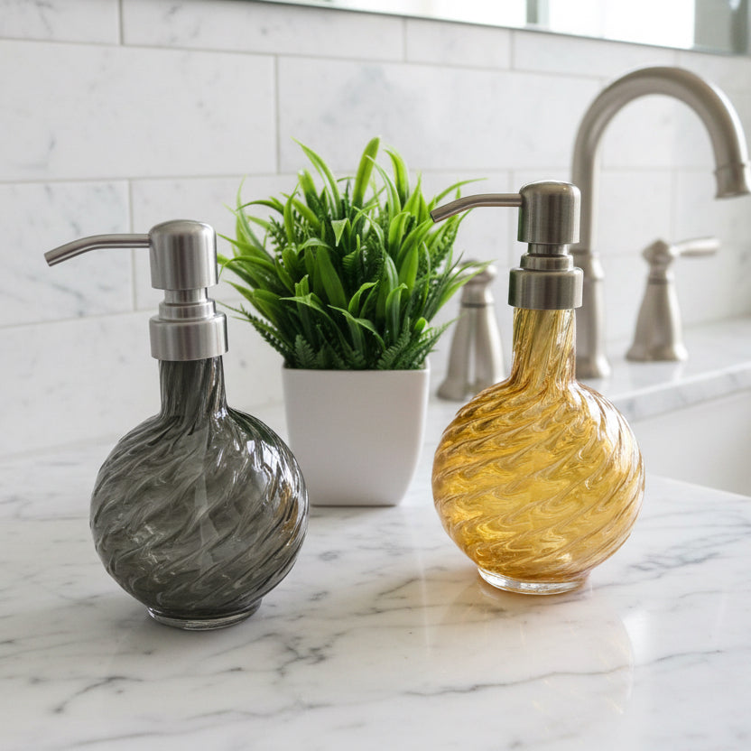 Handblown marbled glass soap dispensers in gray and gold with textured swirl design and metal pumps near a small green plant in a white pot