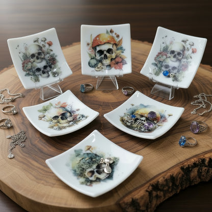 Six small square white ceramic plates with glossy finishes, each featuring a painted skull illustration decorated with colorful mushrooms and botanical elements, displayed on clear acrylic stands against a white background.