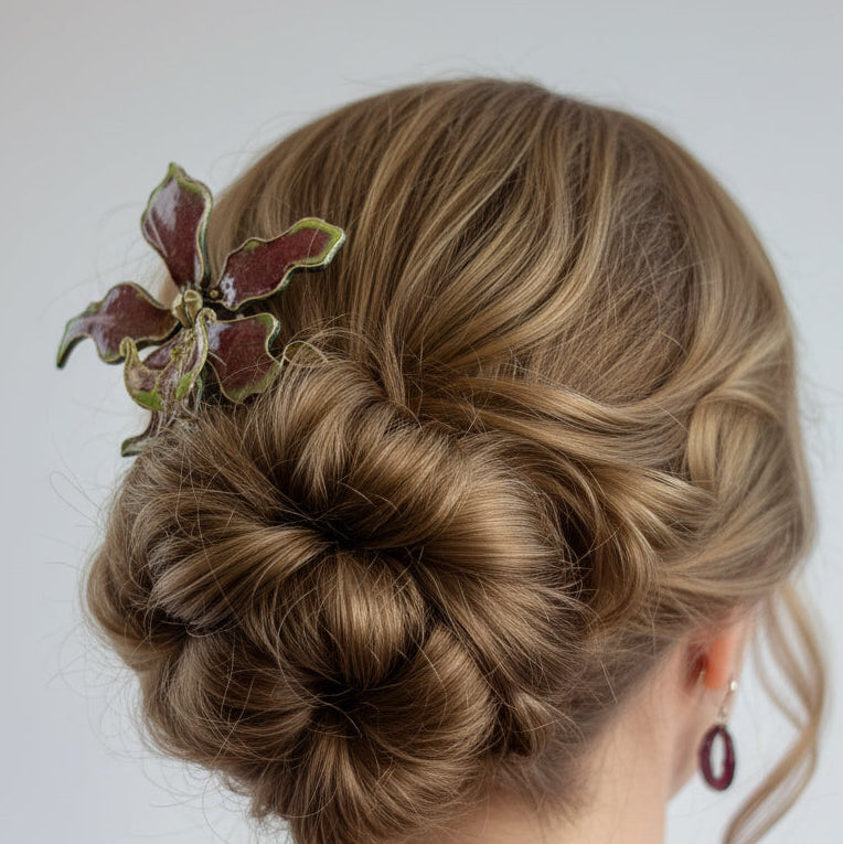 A woman shown from the back wearing an intricate low bun hairstyle with a decorative leaf-shaped hair accessory in burgundy and green, against a plain light gray background.