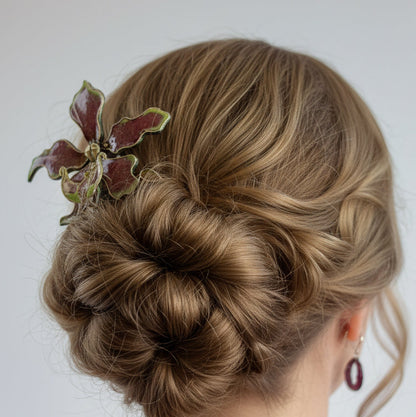 A woman shown from the back wearing an intricate low bun hairstyle with a decorative leaf-shaped hair accessory in burgundy and green, against a plain light gray background.
