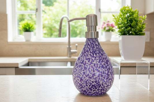 Hyacinth Riverstone hand blown glass soap dispenser with marbled purple and white pattern and stainless steel pump next to green potted plants