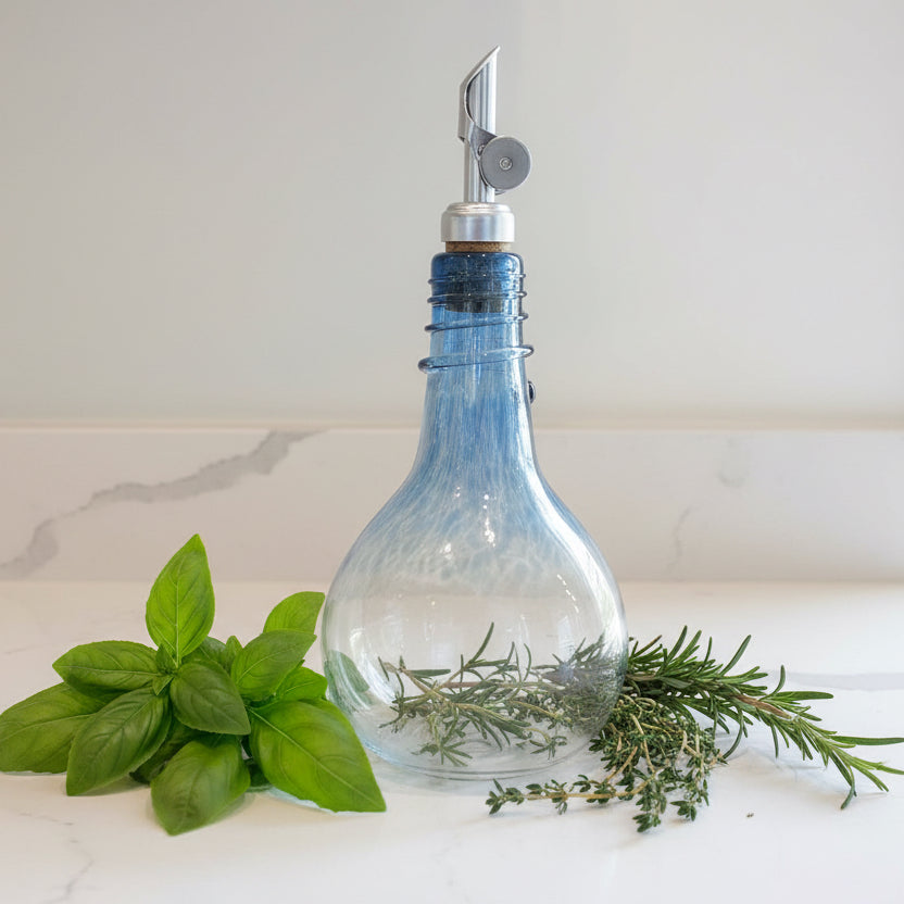 Clear glass Blue Spiral Oil drizzler bottle with a deep blue swirl design and stainless steel spout on a white surface with two green potted plants in white pots in the background