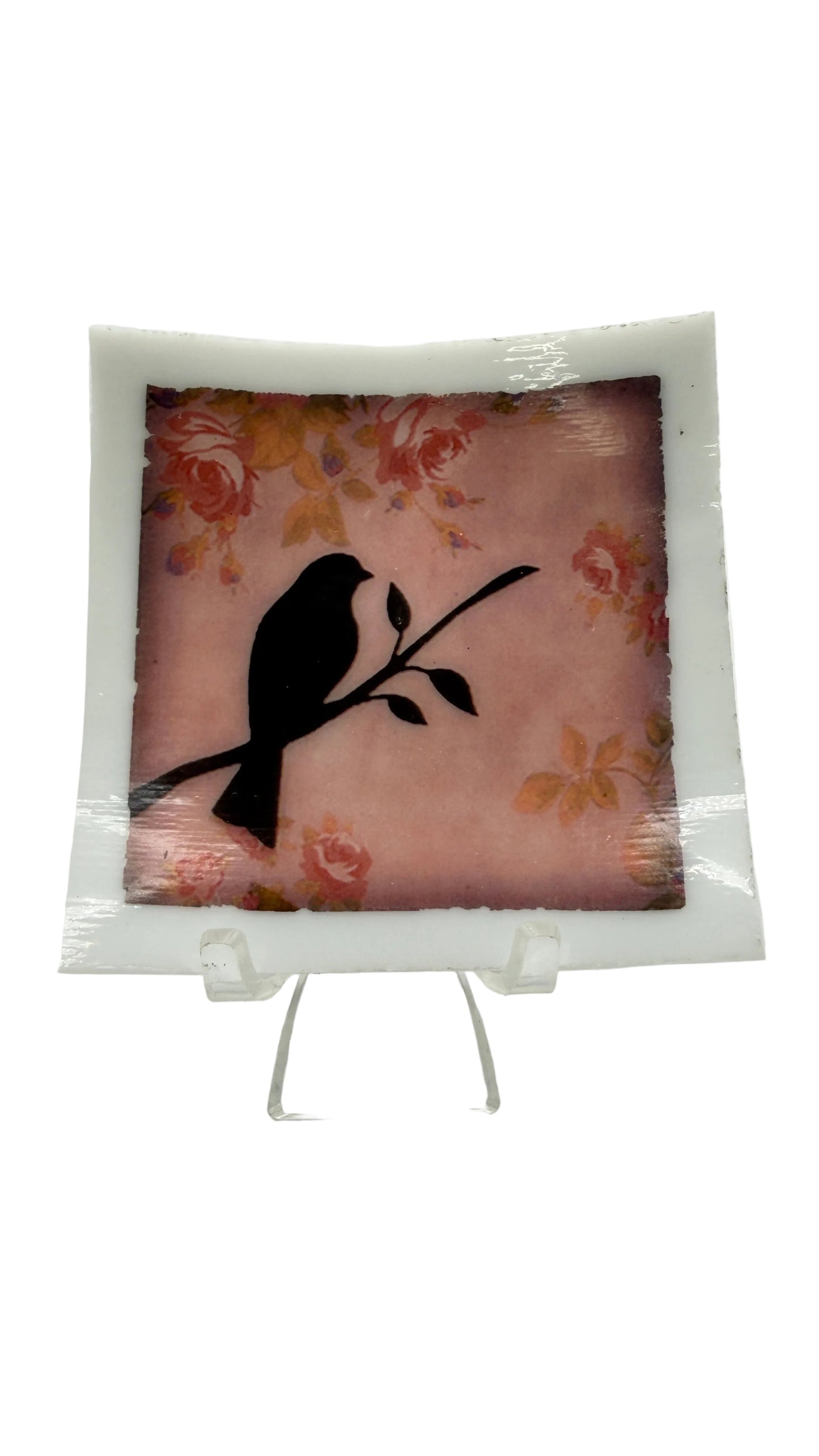 A square decorative glass plate with a white border, featuring a black bird silhouette perched on a branch over a pink floral background, displayed on a clear acrylic stand against a white studio background.