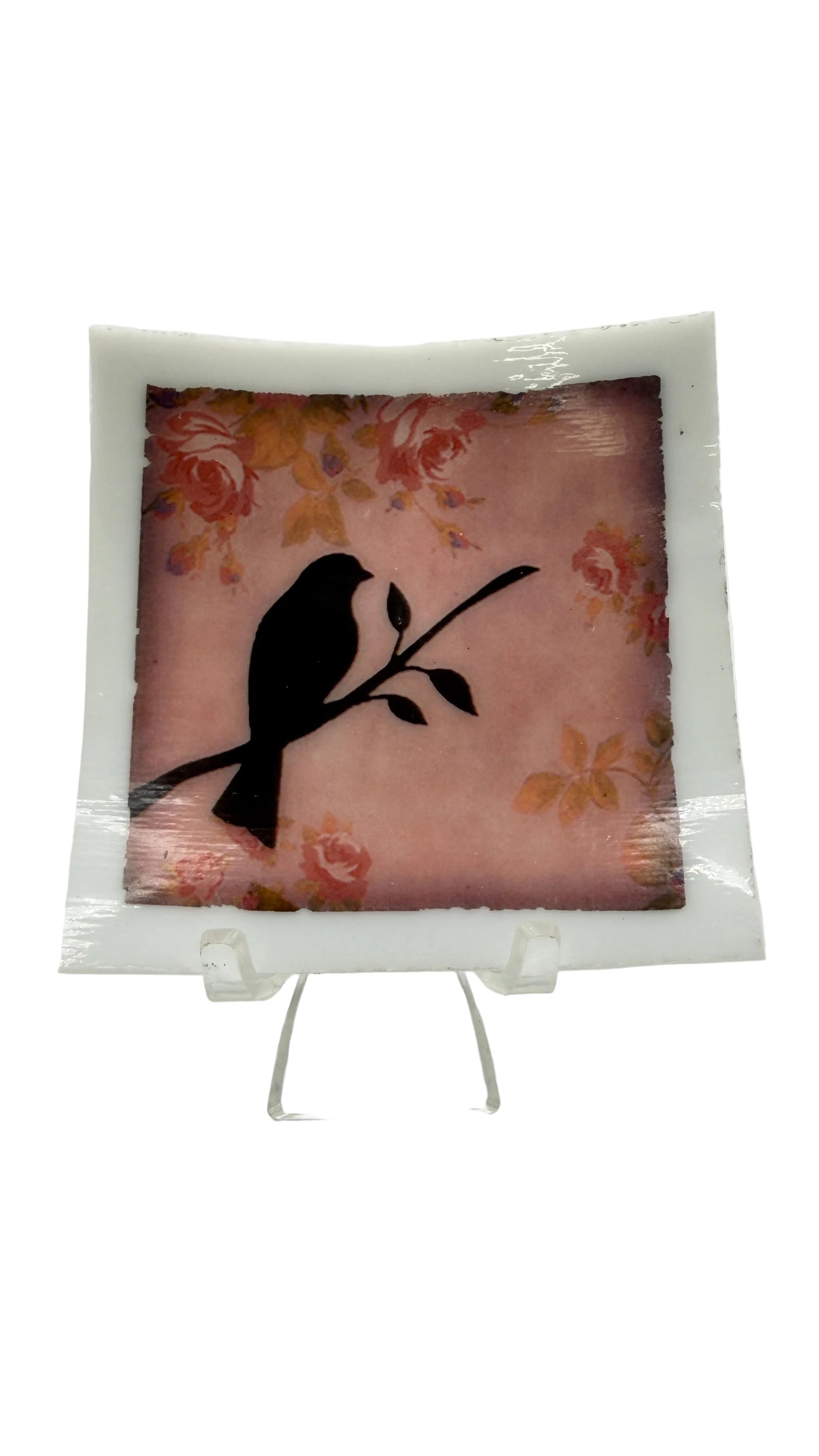 A square decorative glass plate with a white border, featuring a black bird silhouette perched on a branch over a pink floral background, displayed on a clear acrylic stand against a white studio background.