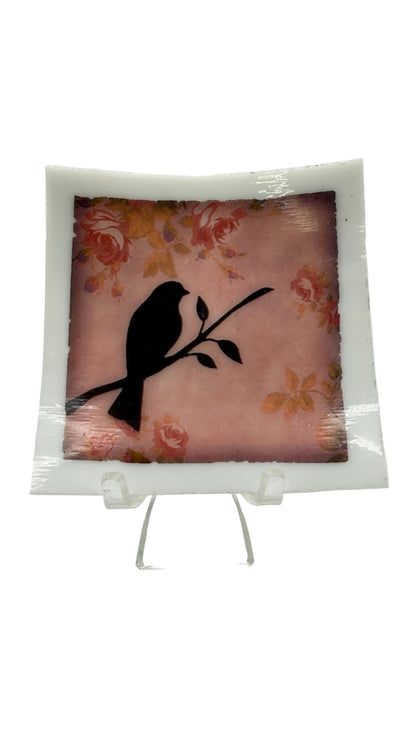 A square decorative glass plate with a white border, featuring a black bird silhouette perched on a branch over a pink floral background, displayed on a clear acrylic stand against a white studio background.