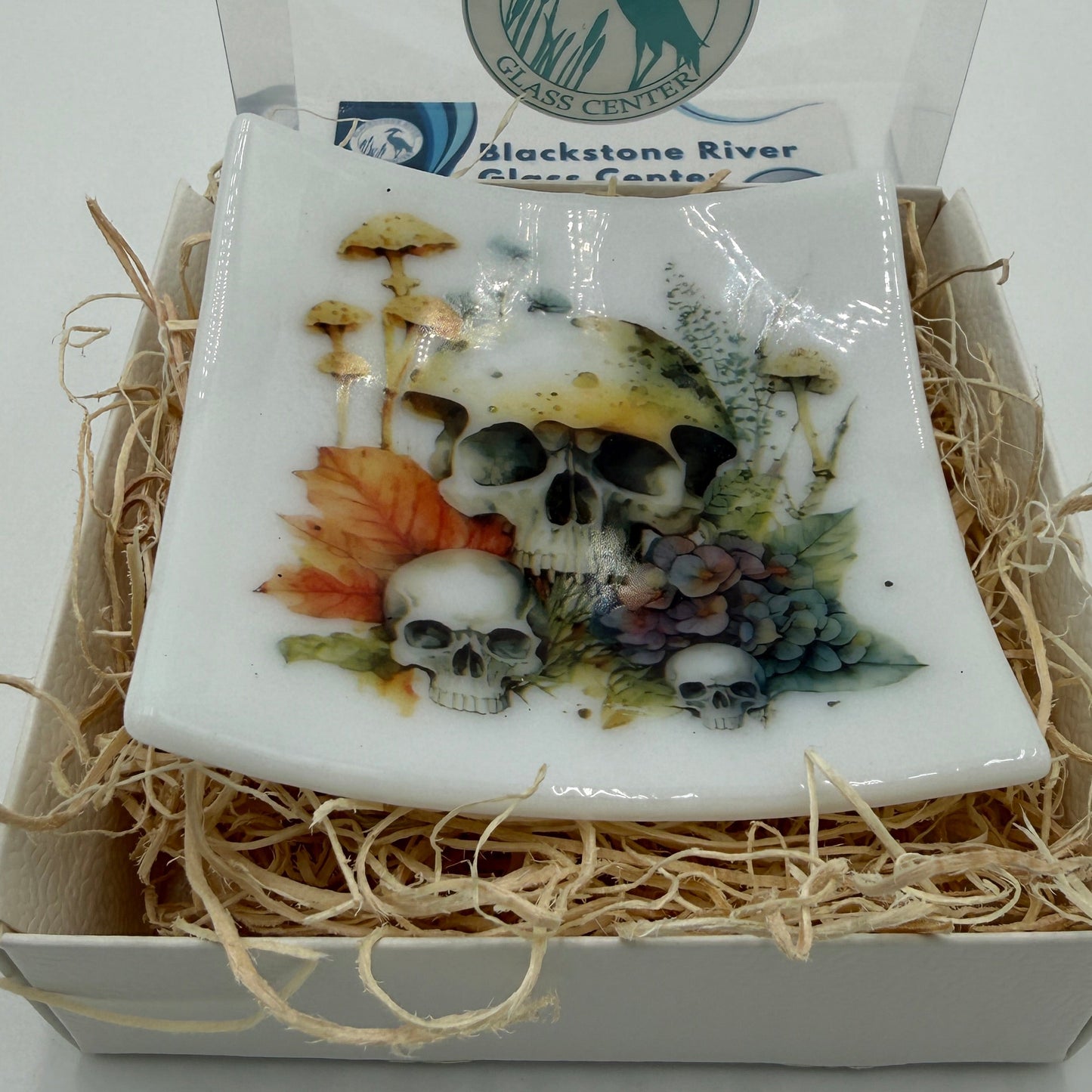 A square white glass  plate featuring a detailed graphic of skulls, mushrooms, autumn leaves, and grapes, presented in a gift box with shredded paper and a clear lid. The lid features the 'Blackstone River Glass  logo.