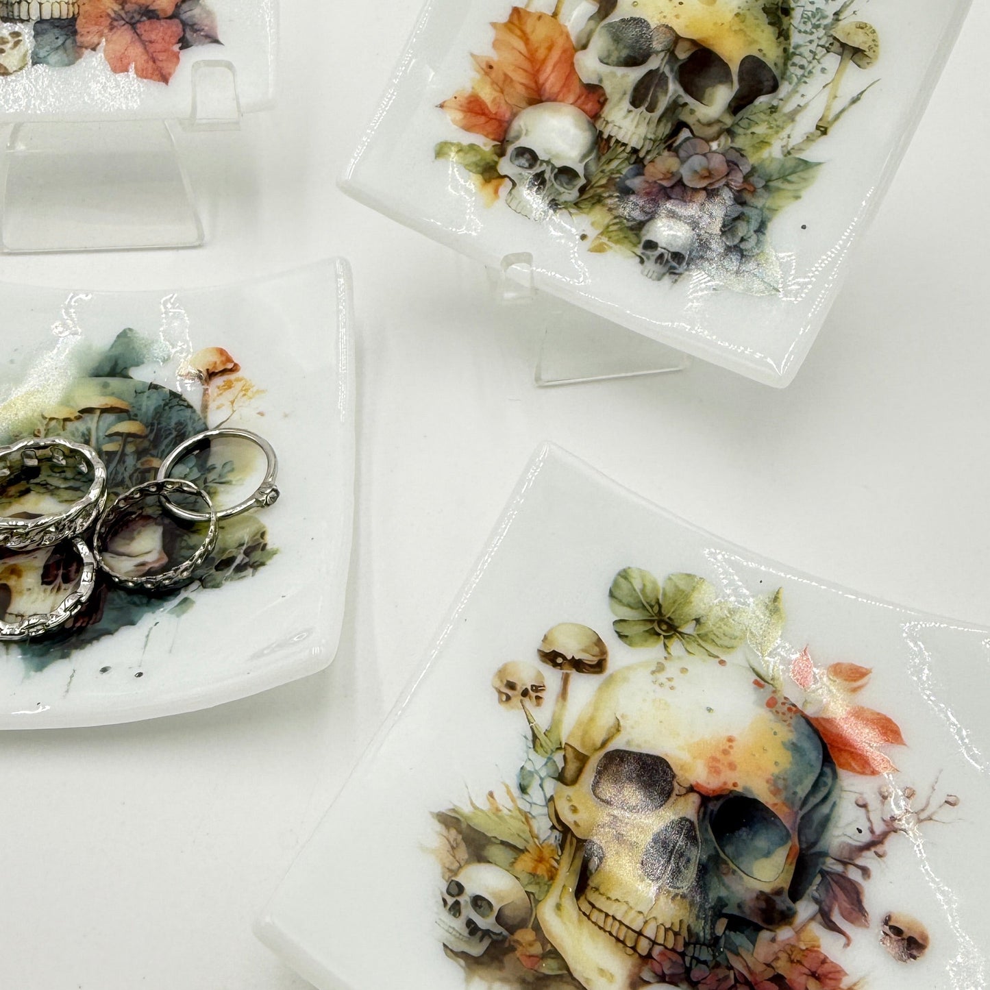 Three square white ceramic plates with rounded corners, each decorated with colorful skull and mushroom illustrations, displayed on clear stands against a white background.
