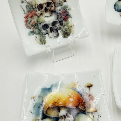White square ceramic plates featuring detailed illustrations of skulls and mushrooms, displayed on small clear stands against a white background.