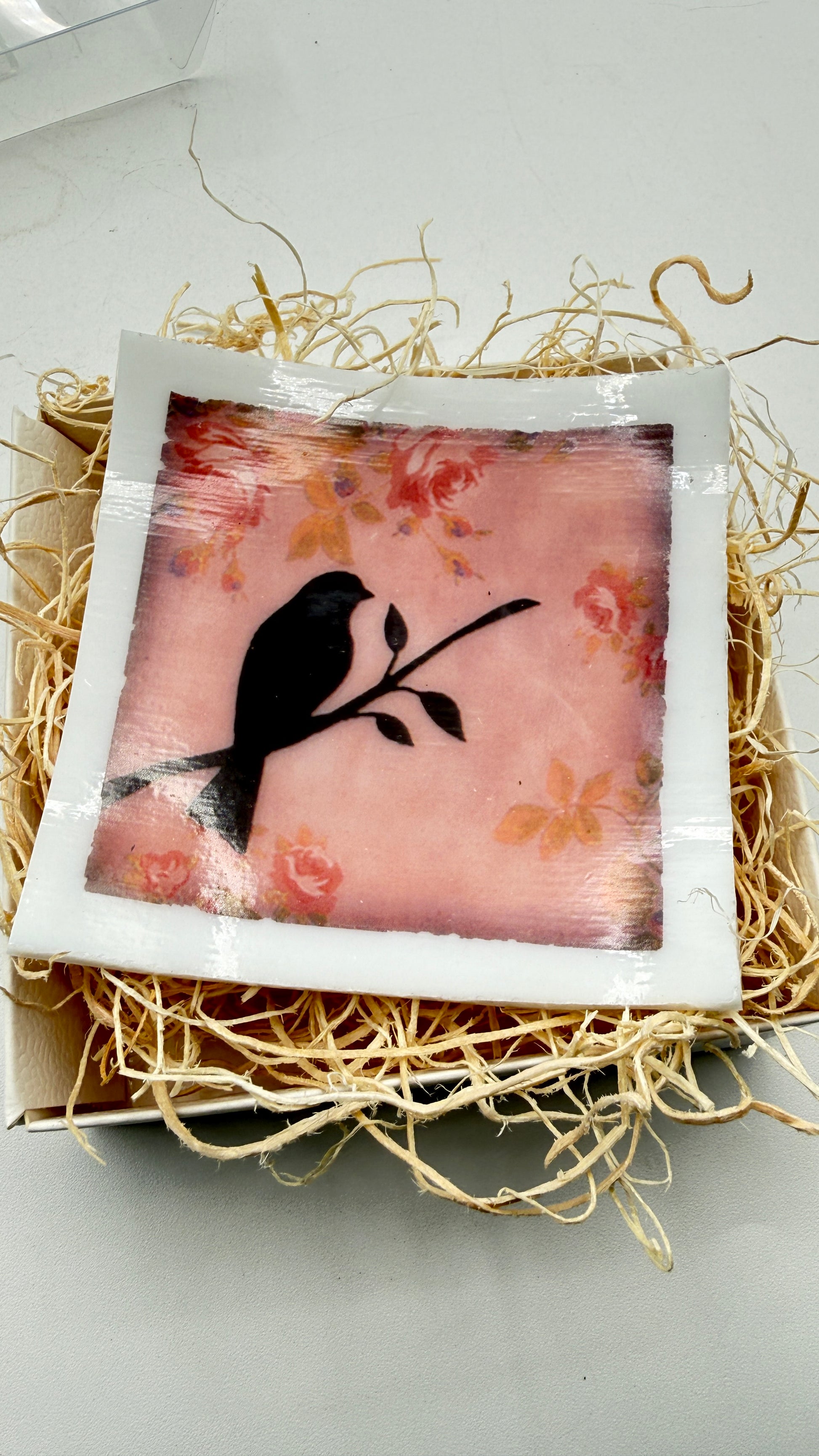 A square decorative plaque or coaster with a white border and a printed design of a black bird perched on a branch against a pink floral background, displayed on a bed of shredded kraft paper inside a box.