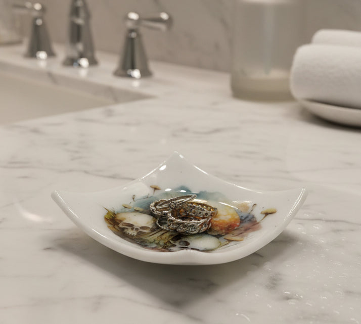 A square white glass ring holder with a fused glass decal of skulls and mushrooms design in the center, holding three silver rings, on a plain white background.