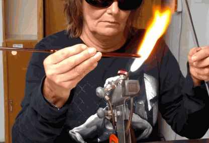 Woman wearing protective glasses shaping molten glass with a bench torch in flameworking studio during a private group glass experience
